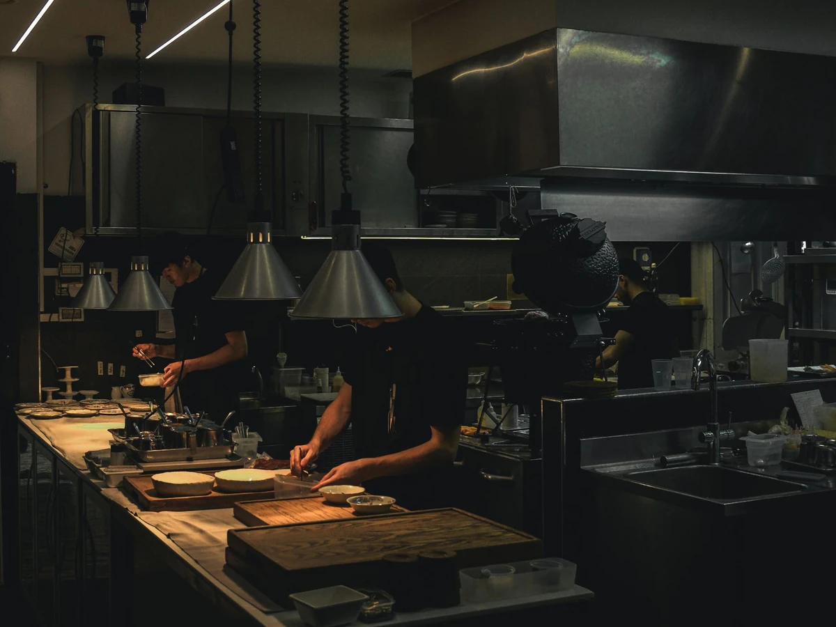 Chefs working the line in a dimly lit professional restaurant kitchen with stainless steel hood and heat lamps