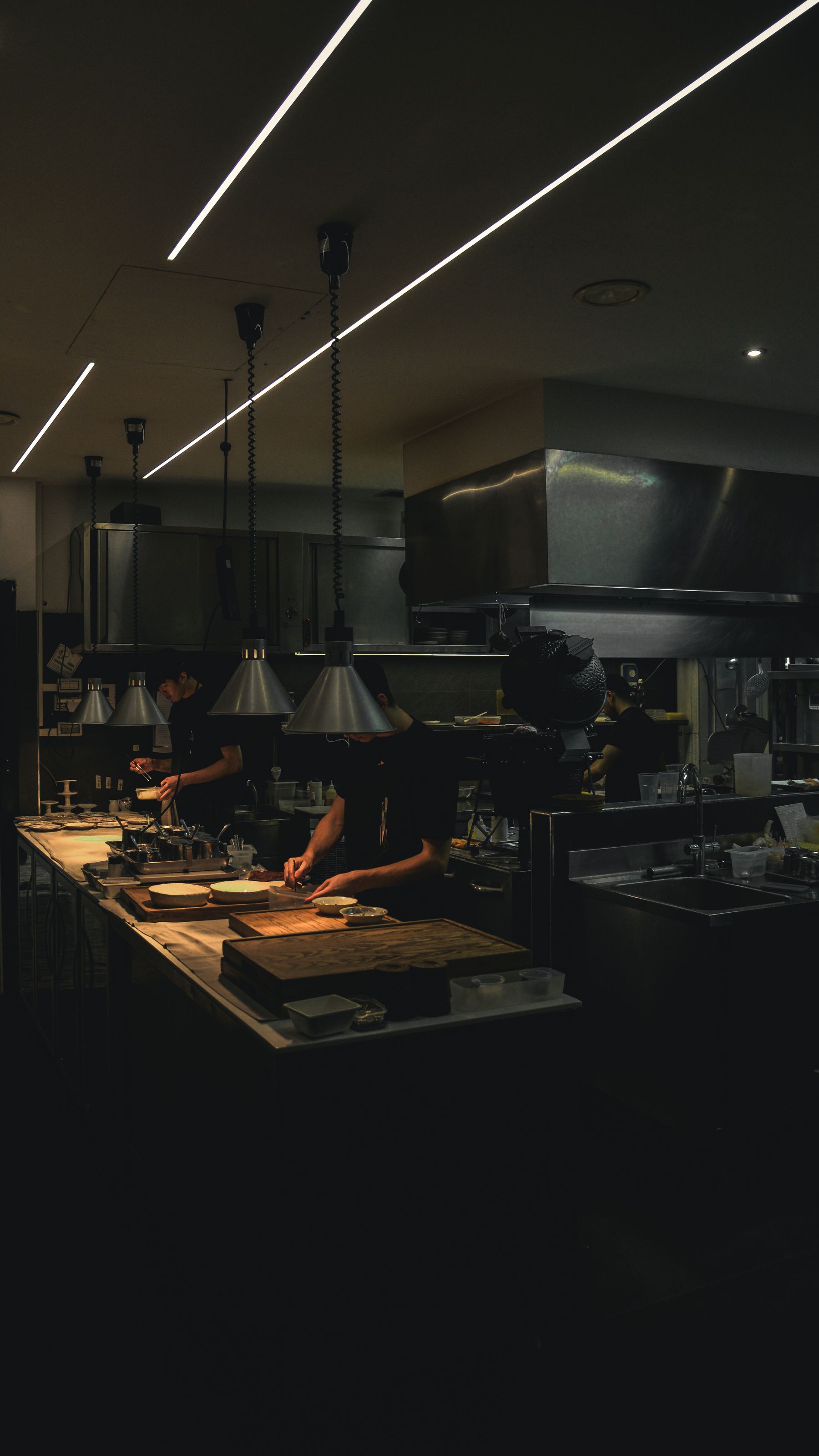 Chefs working the line in a dimly lit professional restaurant kitchen with stainless steel hood and heat lamps